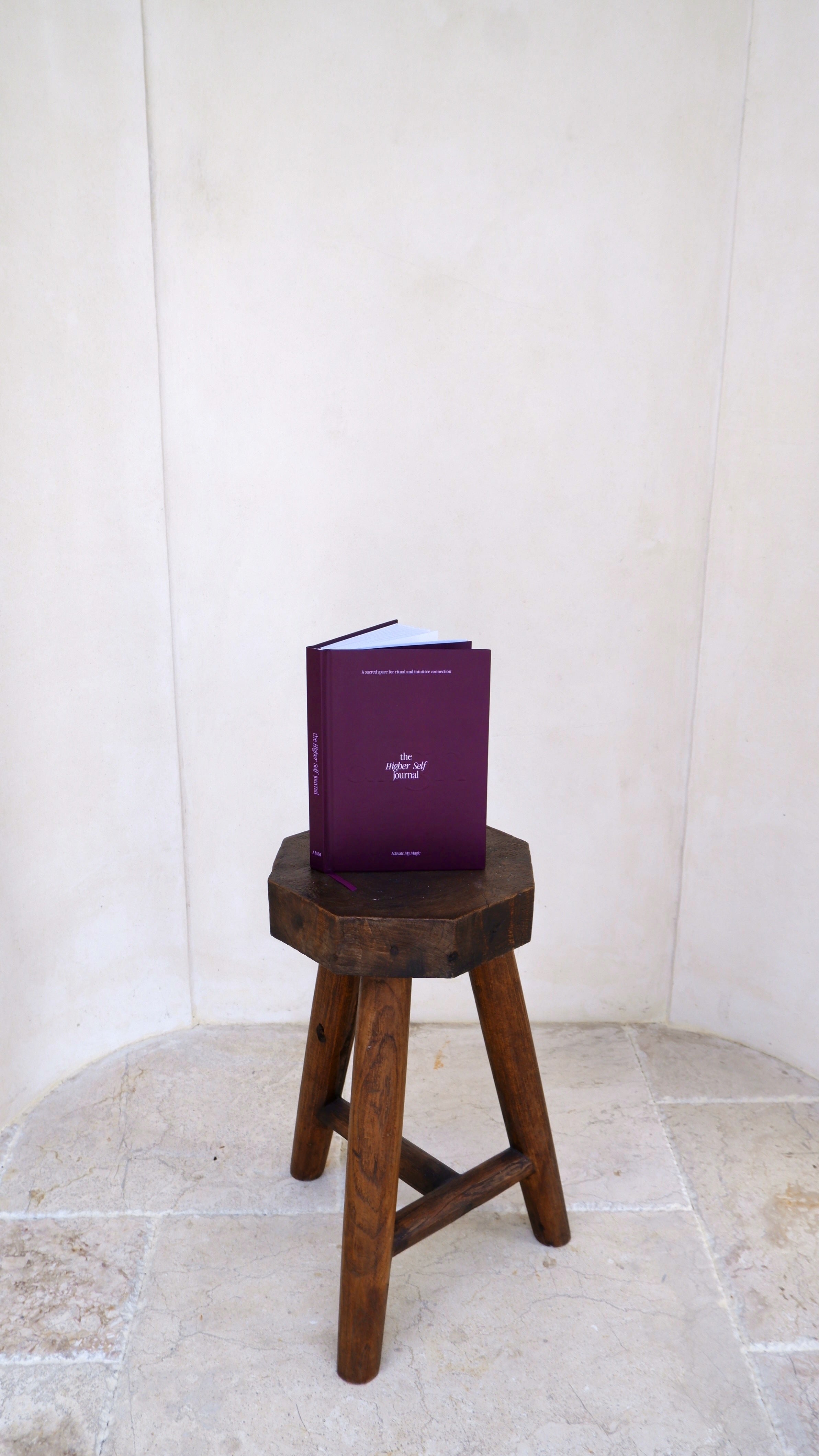A purple book with white text standing upright on a rustic wooden stool against a light beige plaster wall and stone floor.