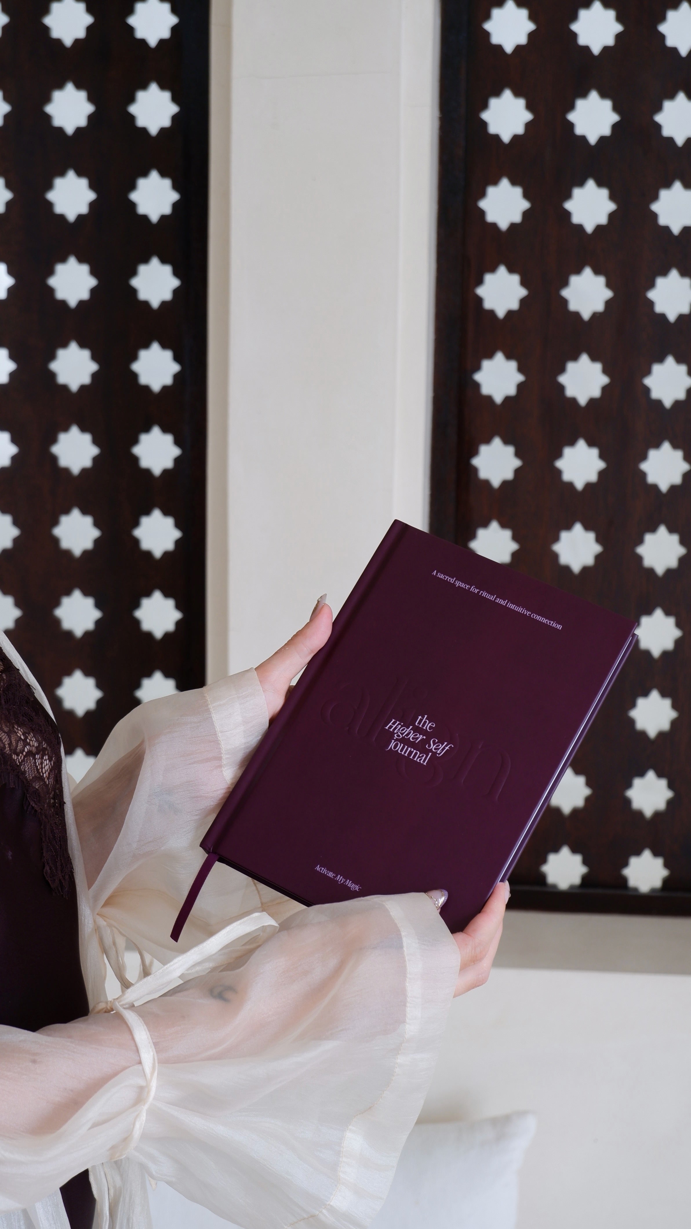 A person in a white sheer kimono holds a dark burgundy hardcover notebook with gold lettering that reads 'the Higher Self journal'. The background features a white wall and dark brown wooden screens with white star patterns.