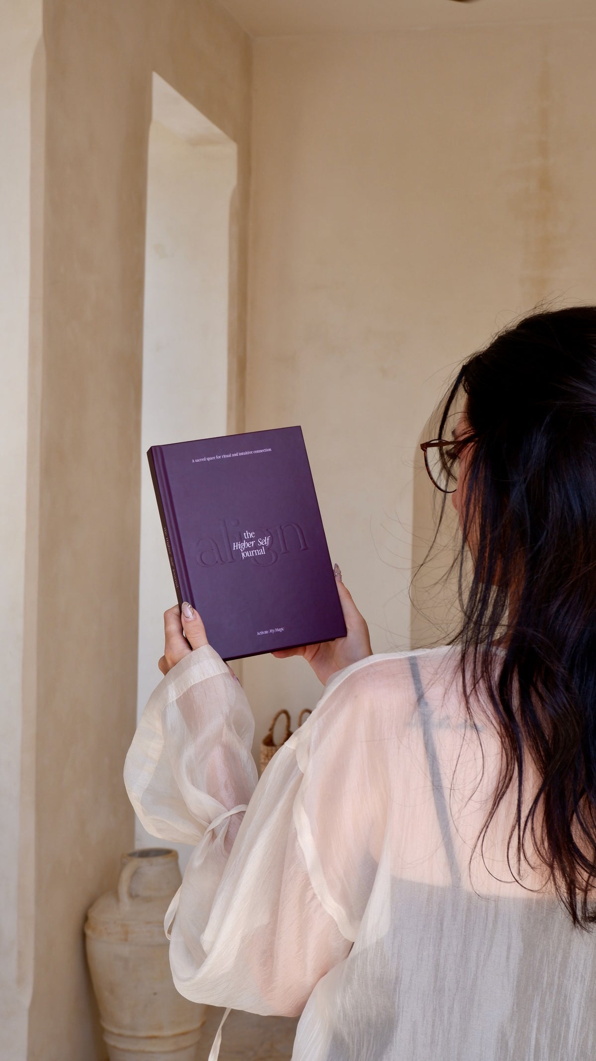 A woman with long dark hair, seen from behind, holding up a purple hardcover journal titled "the higher self journal" against a warm beige wall background.