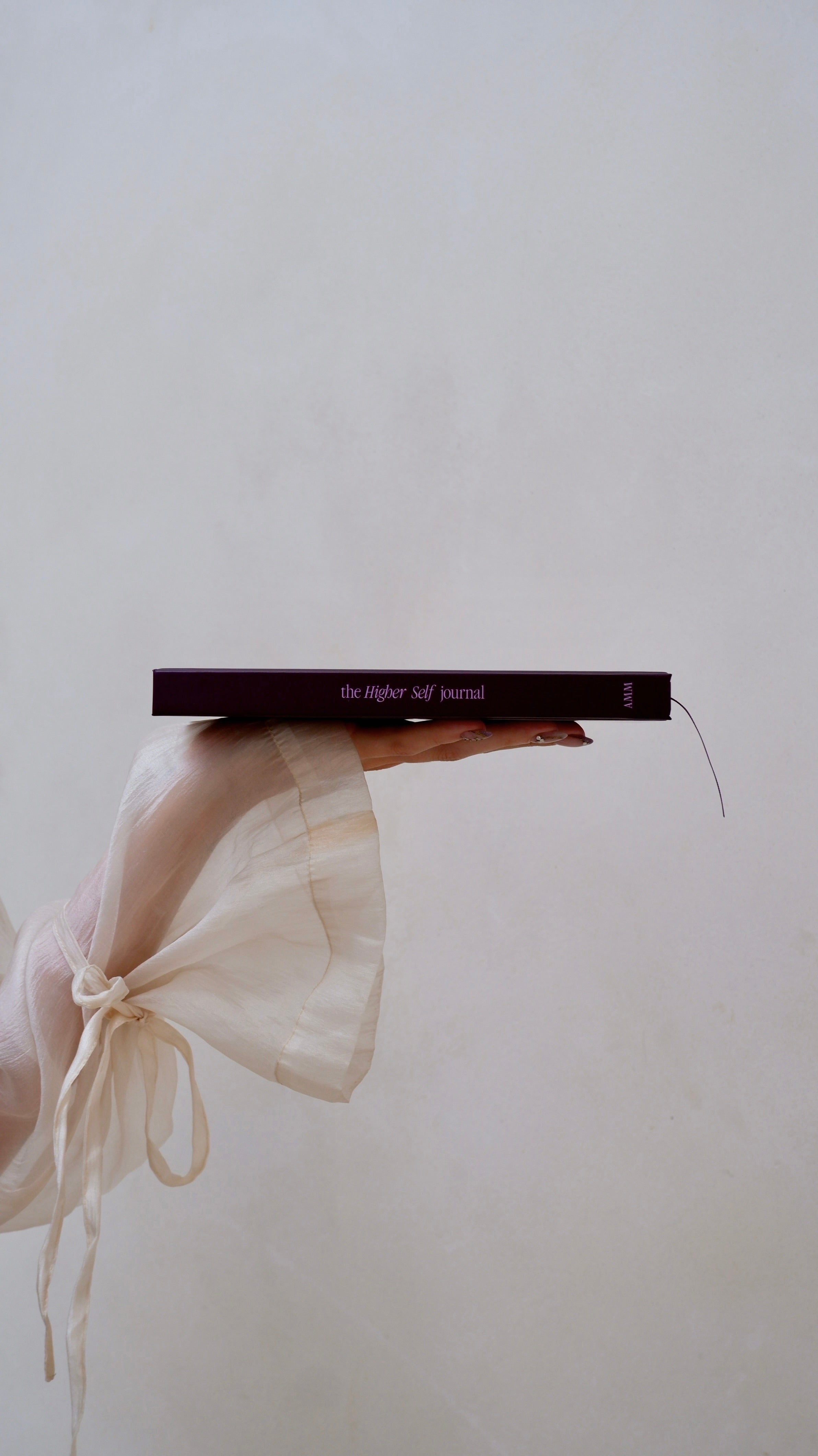 A hand holding a slim dark journal with a sheer cream fabric sleeve tied around the wrist, set against a plain light gray background.